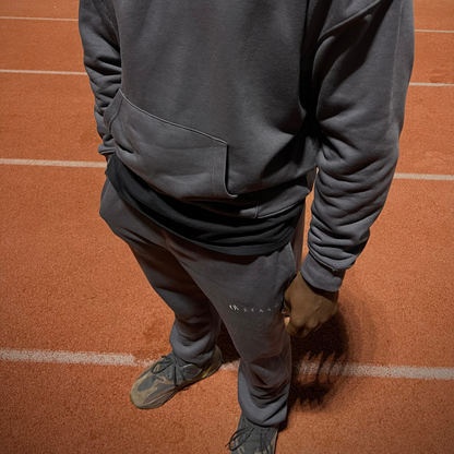 Person wearing a dark gray Ox Season hoodie and matching sweatpants, standing on an outdoor track with a hand in the hoodie pocket. The Ox Season logo is visible on the sweatpants, and the outfit has a comfortable, athletic fit